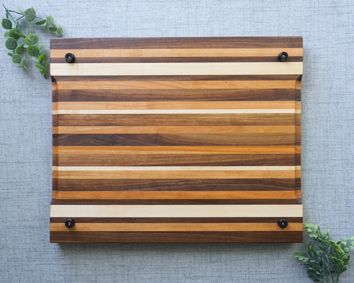 Pattern edge grain cutting board shown on grey table top. Bottom side of cutting board made from domestic hardwoods: black walnut, cherry and hard maple. Board features rubber non slip feet and finger grooves.
