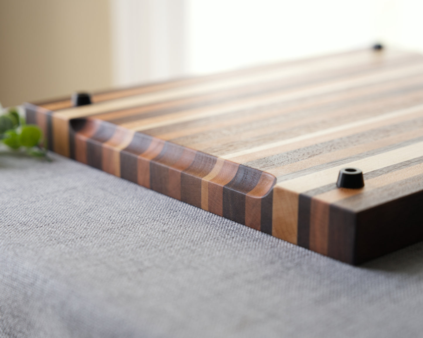 Detail view of a pattern style edge grain cutting board shown on grey table top. Cutting board made from domestic hardwoods: black walnut, cherry and hard maple. Features rubber non slip feet and recessed finger holds.