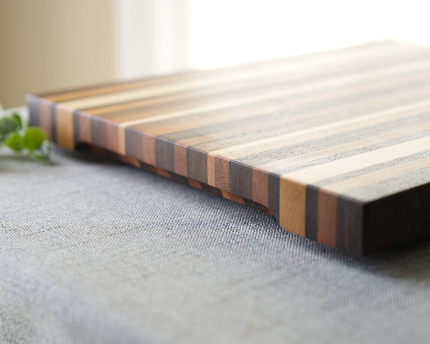 Pattern edge grain cutting board shown on grey table top. Side view of cutting board made from domestic hardwoods: black walnut, cherry and hard maple. Board is set on black rubber feet and features a rounded hand hold on 2 sides.