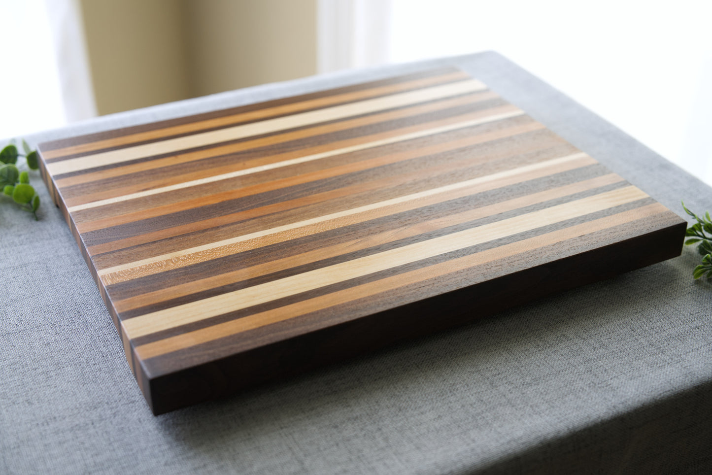 Pattern edge grain cutting board shown on grey table top. Cutting board made from domestic hardwoods: black walnut, cherry and hard maple.