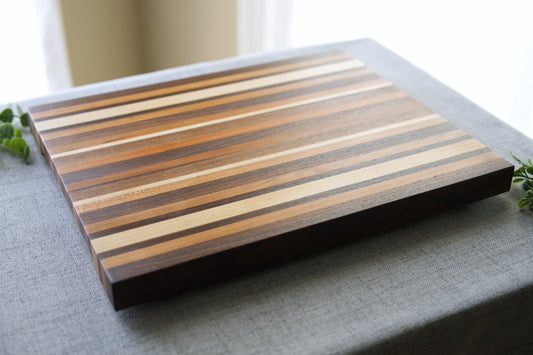 Pattern edge grain cutting board shown on grey table top. Cutting board made from domestic hardwoods: black walnut, cherry and hard maple.